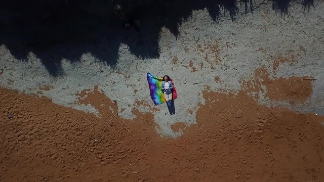 Woman filming herself with a drone at the beach on Gozo island while she is twinking and smiling into the camera as the drone flies up, giving an overview of the golden, orange beach.
