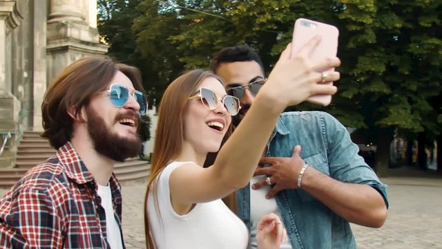Cheerful multiethnic friends in casual clothing and sunglasses smiling and making selfie on smartphone near ancient cathedral