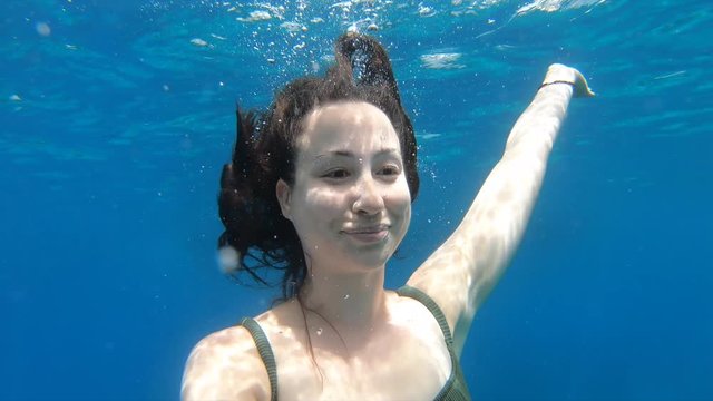 Beautiful, caucasian woman with brown long hair filming herself with a gopro underwater in the clear, blue ocean on comino island in malta.