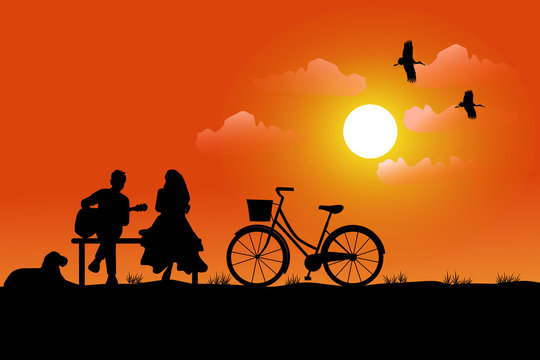 The silhouette of a man playing a guitar for a woman to listen, having a dog and a bicycle beside him. Has a sunset background