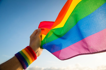Hand with rainbow colors wristband waving gay pride flag waving backlit in the wind against a bright blue sky
