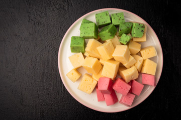 Cheese cubes of green, yellow and red on a plate. Black background. Top view
