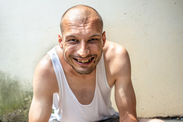 Young skinny anorexic bald positive and happy smiling homeless man sitting on the urban street in the city or town near white wall with big smile, homelessness social documentary concept - obrazy, fototapety, plakaty