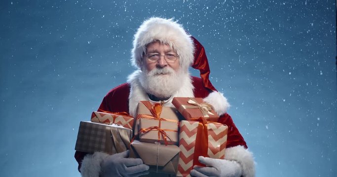 Close up shot of senior bearded man in santa clause outfit carrying gift boxes, isolated on blue background - christmas spirit, holiday mood 4k footage