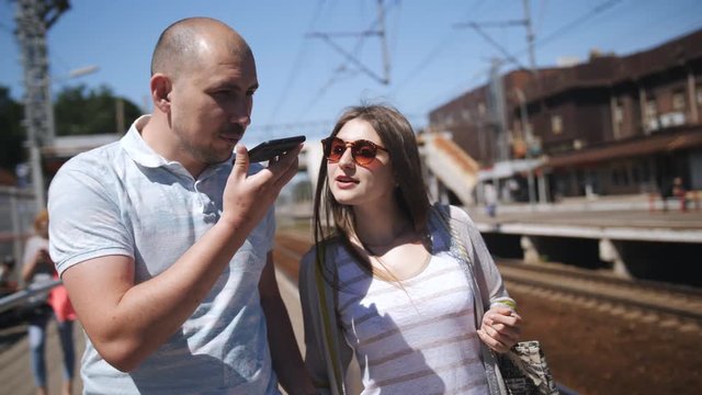 Man and woman are standing at the platform, looking at mobile phones. They are waiting for train arrival, looking at the route in the application on the smartphone.