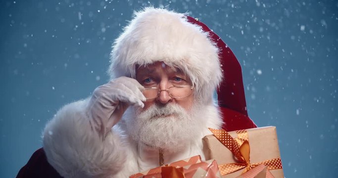 Old bearded man in santa claus clothes taking off his glasses and threatening with his finger, isolated over snowy blue background - christmas spirit concept 4k close up