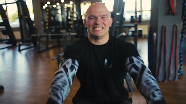bald muscular guy in black dressing breathes deeply doing seated cable rows in gym slow motion closeup