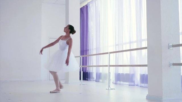 Young girl in white tutu and pointes performances classic ballet dance elements in ballet class. Professional ballet dancer is raising her leg standing on tiptoe and spinning around herself.