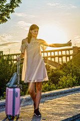 Young beautiful girl standing with a suitcase on a pink background a beautiful sunset.