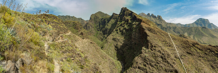 Aerial view of famous Hell gorge in Adeje. Sunny day. Blue sky and clouds above the mountains. Rocky tracking road in dry mountain area. Tenerife, Canary Islands. Wide angle panorama