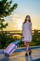 Young beautiful girl standing with a suitcase on a pink background a beautiful sunset.