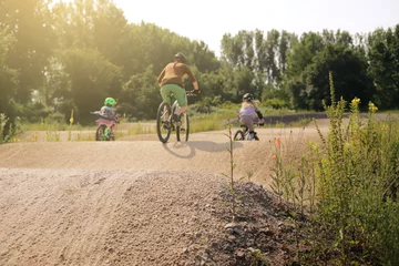 Fototapete Rund mother and two daughters riding bike together on a bicycle dirt track in bright summer light - family lifestyle outdoor activity concept - focus on hill tip in foreground - rider blanked out blurry © R_boe