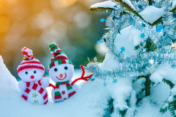 Two small funny toys baby snowman in knitted hats and scarves in deep snow outdoors near pine tree branch. Happy New Year and Merry Christmas greeting card.