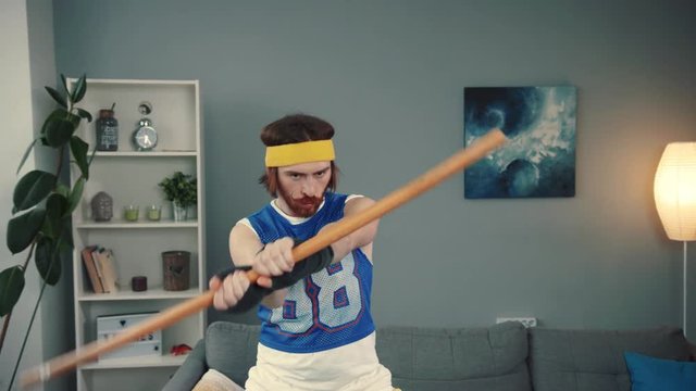 Fun fitness training at home with a wooden stick. Portrait of confident silly sports guy in retro outfit exercising with a stick in funny poses on mat in the living room.