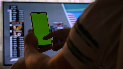 Mockup Green Screen Smartphone. Close Up. Chroma Key Image Of man's Hands Holding Mobile Phone. Against the Background of the TV at home (at the bar) touch screen.