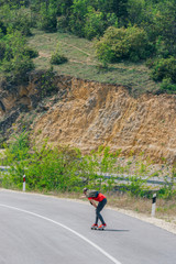 Male caucasian longboarder riding downhill on an empty road doing a speed tuck and grabbing the board while driving the longboard fast. Wearing a red t-shirt green hat and black jeans.