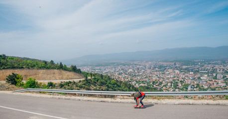 Male hipster wearing a hat and a red shirt is riding a longboard downhill on an open road