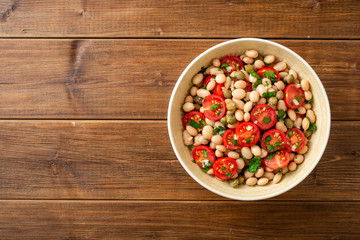 White bean salad with tomatoes, capers, garlic and parsley in bowl on wooden table. Top view. Copy space.