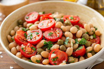 White bean salad with tomatoes, capers, garlic and parsley in bowl on wooden table. Selective focus.