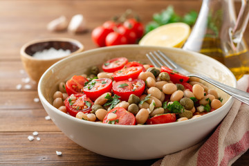 White bean salad with tomatoes, capers, garlic and parsley in bowl on wooden table. Selective focus.