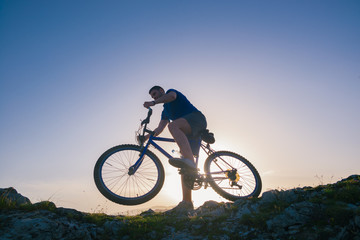 Naklejka na ściany i meble Strong fit male mountain biker performing stunts on rocky terrain on a sunset while wearing a blue shirt and riding a blue bike