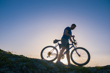Fototapeta na wymiar Strong fit male mountain biker performing stunts on rocky terrain on a sunset while wearing a blue shirt and riding a blue bike