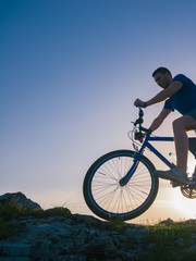 Strong fit male mountain biker performing stunts on rocky terrain on a sunset while wearing a blue shirt and riding a blue bike