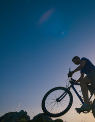 Obraz premium Strong fit male mountain biker performing stunts on rocky terrain on a sunset while wearing a blue shirt and riding a blue bike