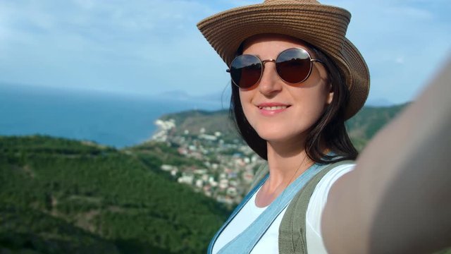 Close-up smiling travel woman in sunglasses and hat taking selfie showing seascape enjoying vacation