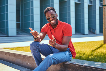 Young happy afro american man in sunglasses is sitting on the grass near skyscraper.