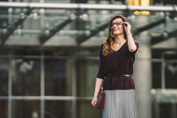 Business woman standing on street against office building. City business woman working. Portrait business woman smiling with glasses. Woman posing outside office building summer business outfit