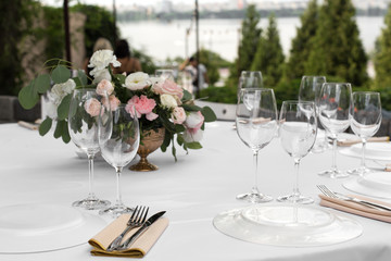 Wedding table setting decorated with fresh flowers in a brass vase. Wedding floristry. Banquet table for guests outdoors with a view of green nature. Bouquet with roses, eustoma and eucalyptus leaves