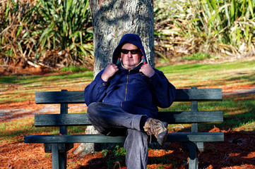 A man sits with is hood up on a seat in the park