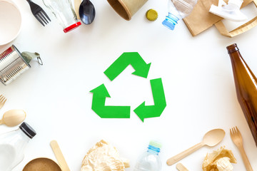 Green recycling sign with waste materials, paper bag and cup, plastic bottles, flatware on white background top view