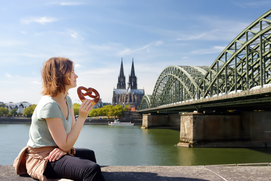 Young woman eats traditional pretzel sitting on embankment of Rhine on background of Cologne Cathedral and Hohenzollern Bridge in Koel, Germany