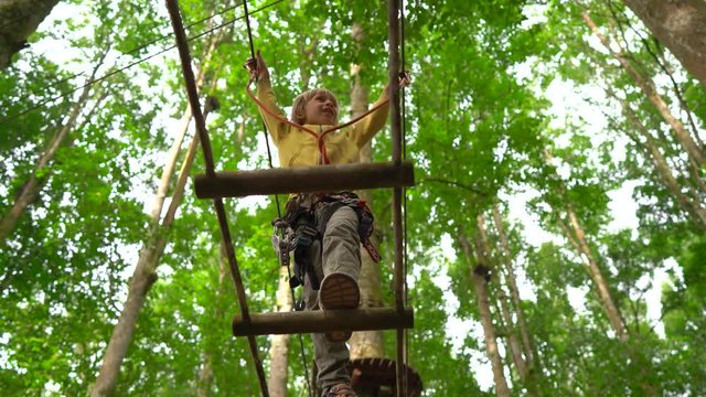 Little boy in a safety harness climbs on a route in treetops in a forest adventure park. He climbs on high rope trail. Outdoor amusement center with climbing activities consisting of zip lines and all