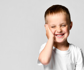 emotional portrait of a little boy in a white t-shirt. The boy looking at the camera, put his palm on his cheek, hesitates and laughs at the one who has been seen.