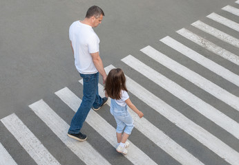 A man and a small child on a zebra crossing trespassing by crossing the street. In the summer on the street kid girl with her father in fashion clothes cross the road.  From top view