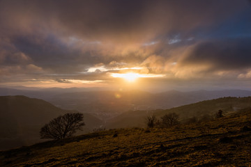 Moody sunset on Monte Cucco (Umbria, Italy), with tree in the foreground and sun filtering through clouds