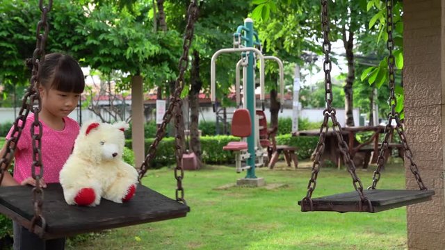 5 years old Asian girl talking and playing with teddy bear on the swing in the park hapily ,summer time.