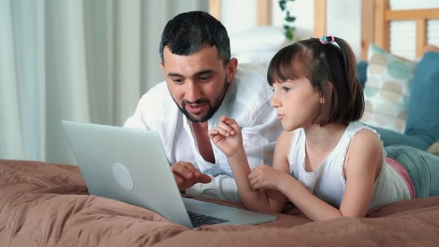 Dad and cute child use laptop lying on bed, plays online game, slow motion