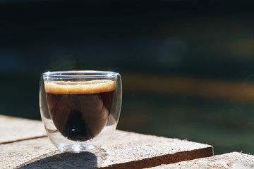 Small transparent double wall glass cup of fresh brewed coffee placed on a natural rough wooden surface in front of a decorative pond