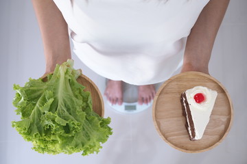 Women weigh themselves on scales and holding vegetables and chocolate cakes. Vegetables are useful to the body and the cake has trans fat. diet concept