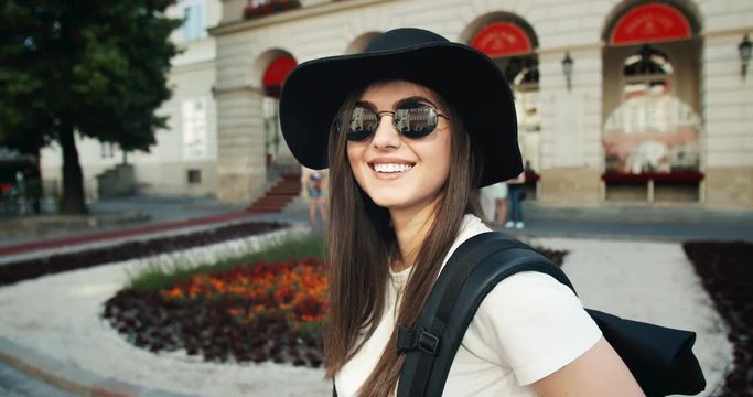 Attractive female tourist in hat and sunglasses standing near city landmark and smiling to camera, portrait