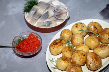 Small young potatoes fried in butter lie in a light plate, next to a saucer with herring, red onion, and a stick with red caviar. Everything is on a gray background. 