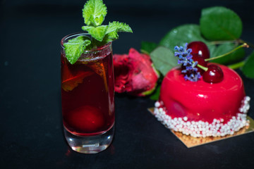 The unlearned cake covered with red icing lies on a dark background, next to a currant compote in a transparent glass, and there is a rose in the background.   