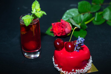 The unlearned cake covered with red icing lies on a dark background, next to a currant compote in a transparent glass, and there is a rose in the background.   