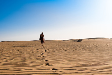 Fototapeta na wymiar young confident woman walking alone on desert sand among dunes on a hot sunny day with clear blue sky