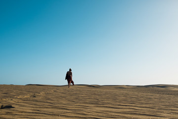 Fototapeta na wymiar young confident woman walking alone barefoot on path in desert sand among dunes on a hot sunny day with clear blue sky
