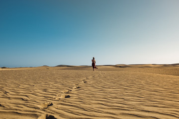 Fototapeta na wymiar evocative young confident woman walking on her own path on the desert sand with red dress in the middlw of dunes on hot summer day with clear blue sky